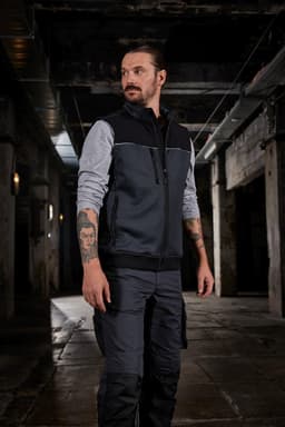 A man wearing a black and grey hybrid workwear vest in an industrial setting, showcasing a sleek design.