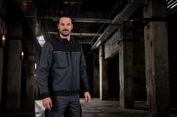 A man stands in an industrial setting wearing a grey and black work jacket with a logo.