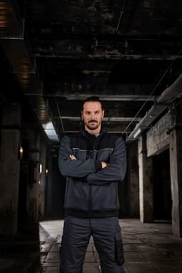 A man wearing a black and grey Hybrid Workwear Jacket stands confidently in an industrial setting.