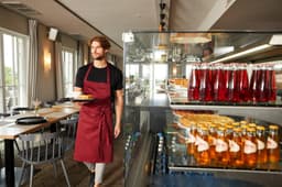 A man wearing a burgundy apron with a bib carries a plate in a modern dining setting, surrounded by drinks in glass containers.