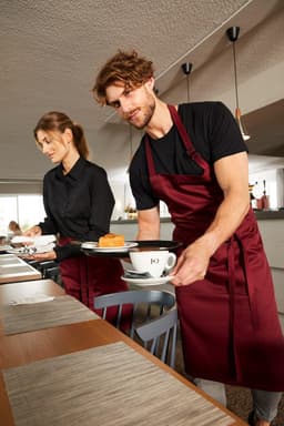 A maroon bib apron is worn by a man serving coffee and dessert in a café setting, alongside a woman in a black shirt.