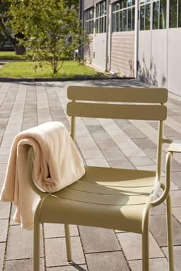 A beige microfibre fleece blanket draped over a light-green chair outdoors, with a paved pathway and greenery in the background.