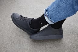 Black sports socks worn with black shoes and blue jeans on a grey surface. The socks have a logo.
