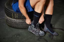 A person is putting on grey shoes while wearing black and grey socks, seated on tyres in a work environment.