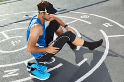 A man in a blue running tank and black leggings stretches on a sports track, wearing a visor and sneakers.
