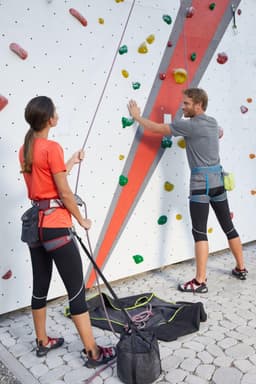 A woman in a red active T-shirt and black leggings stands at a climbing wall, while a man prepares to climb.