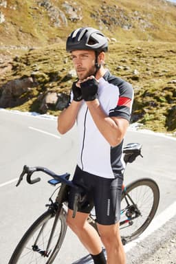 A man in a black and white cycling jersey adjusts his gloves while standing next to a bicycle on a mountain road.