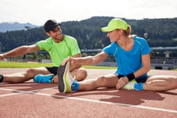 A pair of athletes stretch on a track, wearing vibrant green and blue sports T-shirts and accessories.
