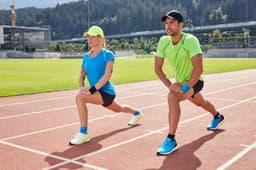 A man in a green sports t-shirt and a woman in a blue sports t-shirt perform lunges on a running track. Both wear caps.