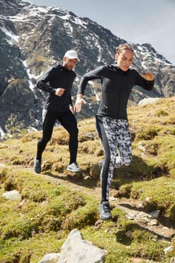A black ladies sports zip hoodie is worn by a woman running through a mountainous landscape, alongside a man in a cap.