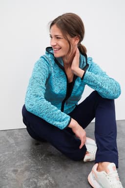 A blue knitted fleece hoodie with a zip and hood, paired with navy pants, worn by a smiling woman sitting on the floor.