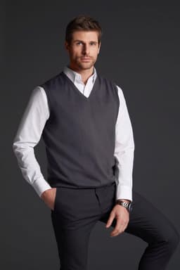 A grey men's v-neck vest is worn over a white shirt, paired with dark trousers. The model is standing with one hand in his pocket.