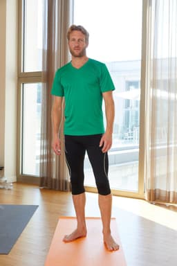 A man stands barefoot on a yoga mat wearing a green active T-shirt and black athletic leggings, with natural light streaming in.