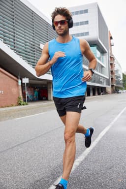 A man in a blue active tank top and black shorts jogs along a city street, wearing sunglasses and headphones.