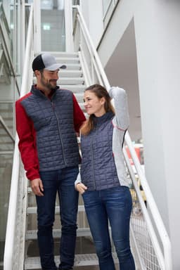 A grey and red knitted hybrid jacket is worn by a man and a woman, standing on a staircase. Both wear casual jeans.