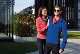 A red ladies fleece jacket and a blue men's fleece jacket are worn by a couple outdoors. Both jackets have a logo.