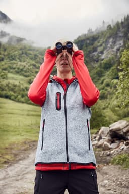 A woman wearing a grey knitted fleece vest with red accents, using binoculars outdoors in a hilly landscape.