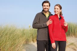 A polar fleece jacket in red, worn by a woman, alongside a man in a grey fleece jacket, with a natural background.