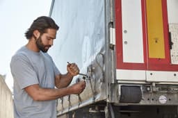 A man in a grey T-shirt uses a wrench on a truck's latch. The setting appears industrial or outdoor.