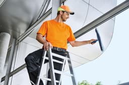 A man in an orange work T-shirt and cap is cleaning windows while standing on a ladder. The setting features modern architecture.