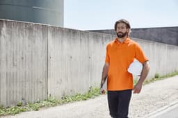 An orange work polo shirt on a man walking outdoors, holding a white helmet and a clipboard.