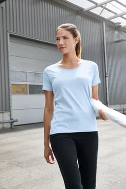 A light blue women's work t-shirt is displayed on a model with black pants, set in an industrial environment.