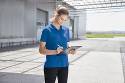 A woman in a blue work polo shirt stands outdoors, holding a tablet and taking notes. The setting features a large, open space.