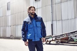 A workwear jacket in navy and blue, featuring a front pocket and logo, worn by a man in an industrial setting.