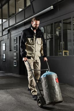 A man in a black and beige workwear jacket is pushing a trolley with a grey cylinder in an industrial setting.