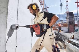 A workwear vest in light beige with dark green accents, worn by a person using a power drill on a construction site.