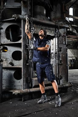 A man in a navy work vest and shorts operates machinery in an industrial setting. The image includes some visible branding.