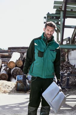 A man stands outdoors wearing a green softshell vest over a black outfit, holding a metal sheet. Logs and machinery are in the background.