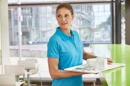 A woman in a bright blue polo shirt carries a tray with cups and a glass in a modern cafe setting.