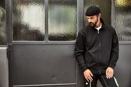 A black workwear sweat jacket worn by a man leaning against a grey door, featuring a logo on the chest.