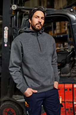 A man in a grey half zip hoodie stands confidently in a warehouse setting with a forklift in the background.
