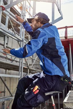 A person in a blue workwear softshell jacket climbs a scaffold with tools visible on their practical pants.