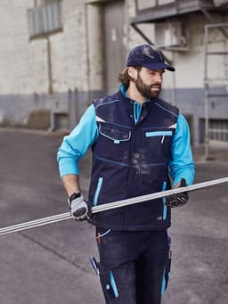 A worker wearing a navy blue softshell padded vest with turquoise accents, holding a metal rod in an outdoor setting.