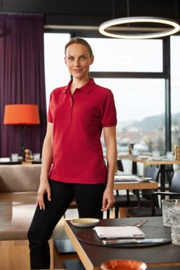 A woman wearing a red polo shirt stands in a stylish indoor setting, showcasing the workwear polo design.