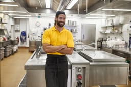 A man in a yellow polo shirt stands confidently in a commercial kitchen, featuring stainless steel equipment and a tidy environment.
