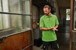A green men's polo shirt is displayed, worn by a man standing near an industrial door. The shirt features a logo.