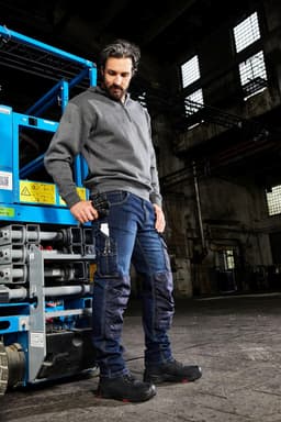 A man wearing grey workwear and blue stretch jeans stands near a worksite with industrial equipment.