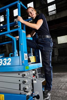 A woman in a black top and navy stretch jeans climbs a blue lift in a warehouse setting.