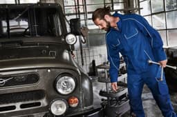 A blue workwear softshell jacket is being worn by a man fixing a vehicle in an industrial setting.