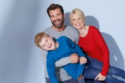 A man in a grey shirt, a woman in a red shirt, and a boy in a blue shirt, smiling together in a casual pose.