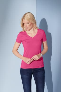 A fitted ladies' t-shirt in pink with a V-neck design, paired with dark blue jeans. The model stands against a light blue background.