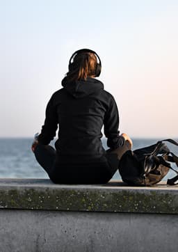 A woman sitting on a ledge wearing a black hoodie and headphones, with a bag beside her, overlooking the water.