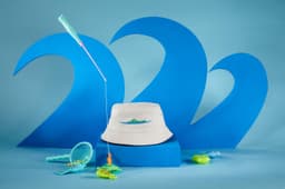 A white fisherman piping hat displayed against blue wave backdrops, surrounded by fishing toys in turquoise and green.