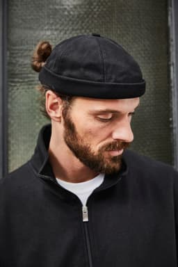 A black chef cap is worn by a man with a beard and a bun, featuring a branded label.