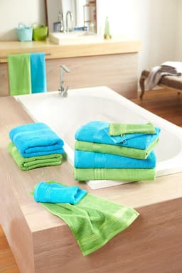 A stack of blue and green hand towels on a wooden surface near a bathtub, with more towels hanging nearby.