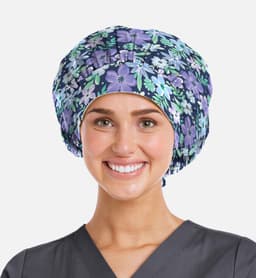 A printed unisex bouffant cap in purple with floral patterns, worn by a smiling individual in a grey scrub top.